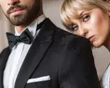 Elegancki mężczyzna w czarnym smokingu z muchą i białą koszulą. Obok niego opiera się kobieta w białej sukni. Idealny przykład, jaka koszula do muchy.