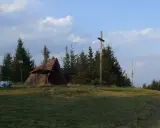 Drewniana wiata, krzyż i kosz na śmieci na tle drzew i nieba.
