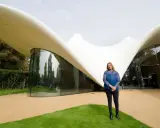 Zaha Hadid steht vor einem ihrer ikonischen zaha hadid bauwerke, einem geschwungenen, weißen Dach mit Glasfront.