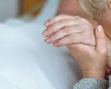 Dłoń osoby starszej trzymająca inną dłoń, na tle białej pościeli.