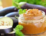 Un bocal de le vrai caviar d'aubergine, garni de persil frais, avec des aubergines entières et tranchées en arrière-plan.