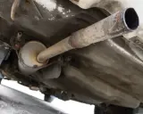 Vista inferior de un coche, mostrando el sistema de escape con un silenciador y tuberías oxidadas. El colector de escape es una parte crucial del sistema.