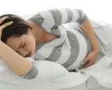 Schwangere Frau liegt auf dem Bett, hält sich den Kopf und fragt sich: "Vorwehen, wie lange noch bis zur Geburt?