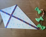 Bastelanleitung mit Bildern: Ein selbstgebastelter Drachen aus Papier mit Schneeflocken und buntem Klebeband, geschmückt mit grünen Schleifen und Holzperlen.