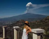 Frau mit Hut blickt auf den rauchenden sizilien vulkan. Die Landschaft ist grün und bergig.