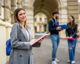 Uśmiechnięta studentka z notatnikiem w ręku, przygotowująca się do studiów 2 stopnia. W tle para studentów.