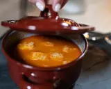 Una mano levanta la tapa de una cazuela de barro con una deliciosa comida típica de Ávila, un guiso naranja con trozos blancos.