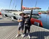 Deux jeunes hommes posent devant un bateau de course prêt pour une traversée atlantique. Le bateau est long, gris et rouge, avec des voiles partiellement visibles.