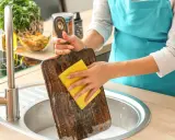 Frau reinigt ein Holz-Schneidebrett mit einem gelben Schwamm in der Spüle. Backpulver hilft, hartnäckige Flecken zu entfernen.