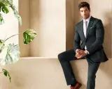 A man in a sharp green suit and black shirt poses confidently next to a lush plant.