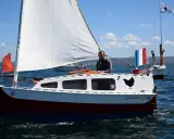 Un homme sourit sur son bateau nommé "Baluchon". Voiles blanches, coque rouge et bleue, drapeau français.