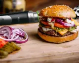 Pyszny burger z serem, warzywami i domowym sosem do burgera. Obok pokrojona cebula i pomidor.