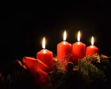 Fyra röda adventsljus brinner i mörkret, en del av en julkrans med granris och röda band. Detta är en visuell representation av vad betyder advent.