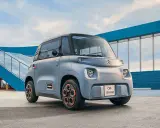 Un cuadriciclo pesado Citroën Ami gris con ruedas naranjas, aparcado frente a una estructura azul y una rampa.