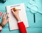 Ręka z czerwoną hybrydą pisze na kartce "Wakacyjny budżet". Obok leżą euro, monety, mapa, lupa i walizka. Czas zacząć planować wakacje za ile!
