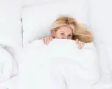 Uśmiechnięta blondynka ukrywa się pod białą kołdrą. Czy to znak, że zastanawia się, jak prać kołdrę?
