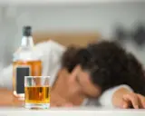 Szklanka z alkoholem i butelka na stole, osoba śpiąca w tle.