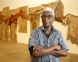 A distinguished man with white hair and glasses, arms crossed, stands before a wall adorned with shimmering, textured artworks, a testament to the enduring legacy of famous artists today.
