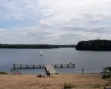 Spokojny dzień na plaży nad jeziorem, żaglówka na wodzie, drewniane molo. Pojezierze Brodnickie zaprasza do wypoczynku.