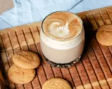 Ein Glas Flat White mit Latte Art neben Keksen auf einem Holztablett. Der Unterschied zwischen Flat White und Cappuccino ist hier deutlich sichtbar.