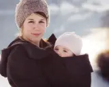 Kvinna med bebis i bärsele, båda med mössor på. Vinterlandskap i bakgrunden.
