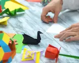 Ręce składają papier, tworząc origami: czarnego łabędzia, czerwoną różę i kolorowe figury. Pokazuje, co można zrobić z papieru.