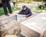 Pracownicy wykonują ekshumację, wyciągając biały worek z grobu. Obok leży drewniana trumna.
