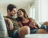 Pareja sonriente en el sofá, compartiendo un momento íntimo. Ella le muestra algo en su móvil, una forma de como expresar sentimientos.
