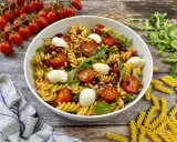 Ein köstlicher Pesto-Salat mit getrockneten Tomaten, Mozzarella-Kugeln und Pinienkernen, serviert in einer weißen Schüssel.