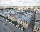 Widok z góry na Rynek Główny w Krakowie. Tłumy ludzi spacerują, a dorożki czekają na turystów. W tle widać zabytkowe kamienice i rozległą panoramę miasta.