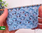 Manos tejiendo un patrón de puntos calados a dos agujas utilisima en hilo azul claro. El tejido muestra un diseño intrincado con agujeros y trenzas.
