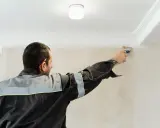 Mężczyzna w roboczym ubraniu maluje pędzlem ozdobny pasek pod sufitem.