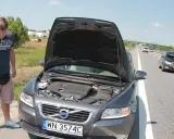 Mężczyzna zmartwiony przy otwartej masce Volvo, prawdopodobnie przegrzany silnik. W tle droga i pola.