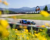 Un coche de F1 azul compite en un circuito soleado, con flores amarillas en primer plano. ¡Un gran resultado f1 hoy!