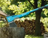 Une hachette bleue et grise est plantée dans un billot, prête à couper du bois. Une pile de bois est visible en arrière-plan.