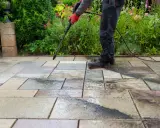 Persona con guantes rojos usa hidrolimpiadora para limpiar suelo terraza de baldosas, rodeado de vegetación y flores.