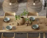 Drewniany stół w jadalni w stylu boho, nakryty do posiłku. Wazon z gałązkami i liśćmi, misy, kieliszki i plecione lampy tworzą przytulną atmosferę.