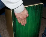 Dłoń z obrączką na palcu spoczywa na zielonym pudle perkusyjnym.
