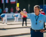 Arbitre de pétanque brandit un carton jaune, rappelant les nouvelles règles. Des joueurs se penchent sur le terrain.