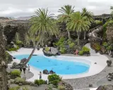 Un oasis de palmeras y cactus rodea una piscina azul en Lanzarote. Ideal para excursiones en Lanzarote con niños, este lugar ofrece un paisaje volcánico único.