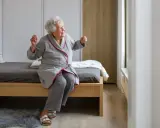 Uśmiechnięta starsza pani w szlafroku siedzi na łóżku, zastanawiając się, jaki wybrać pokój w sanatorium.