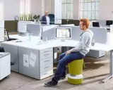 Un espace de travail open space moderne avec des bureaux modulables, des plantes vertes et des sièges colorés. Deux personnes discutent, une troisième travaille sur son ordinateur.