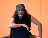 Mauro Corona, con bandana e barba grigia, seduto su una sedia. Immagina i suoi libri in ordine cronologico.