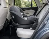 Silla de coche para bebé, instalada a favor de la marcha, con base giratoria y tapicería gris sobre asientos de cuero blanco en el interior de un vehículo.