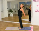 Frau dehnt ihre Brust, Hände hinter dem Rücken verschränkt, in einem hellen Raum mit Spiegeln.