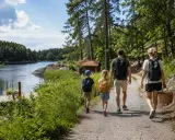 Familj på utflykt Stockholm, vandrar längs en sjö med ryggsäckar och picknickkorg.