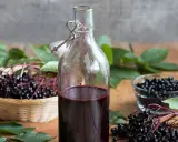 Flasche mit dunklem Saft, daneben Körbchen und Schale mit Holunderbeeren. Achtung: Holunderbeeren giftig!