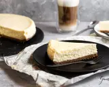 Ein Stück New York Cheesecake auf schwarzer Platte, daneben ein ganzer Kuchen und ein Latte Macchiato.