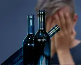 Trzy butelki alkoholu na pierwszym planie, w tle osoba zakrywająca twarz. Symbol walki z nałogiem, nadzieja na odwyk alkoholowy.