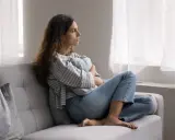 Kobieta siedzi na kanapie, przytulając poduszkę. Zamyślona patrzy przez okno. To pokazuje, po co światu psychoterapia – by odnaleźć spokój.